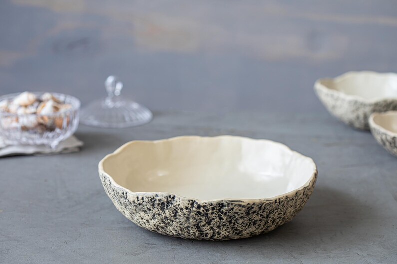 May include: A white ceramic bowl with a speckled black and gray glaze. The bowl has a wide, shallow shape and a slightly rippled edge.