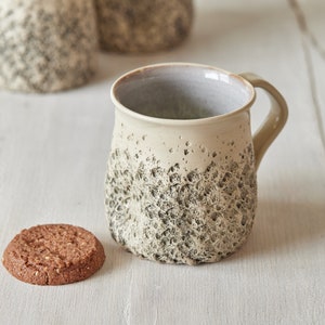 May include: A beige ceramic mug with a textured, speckled surface and a light gray interior. The mug has a rounded body and a handle. A chocolate chip cookie is sitting on a white wooden surface next to the mug.