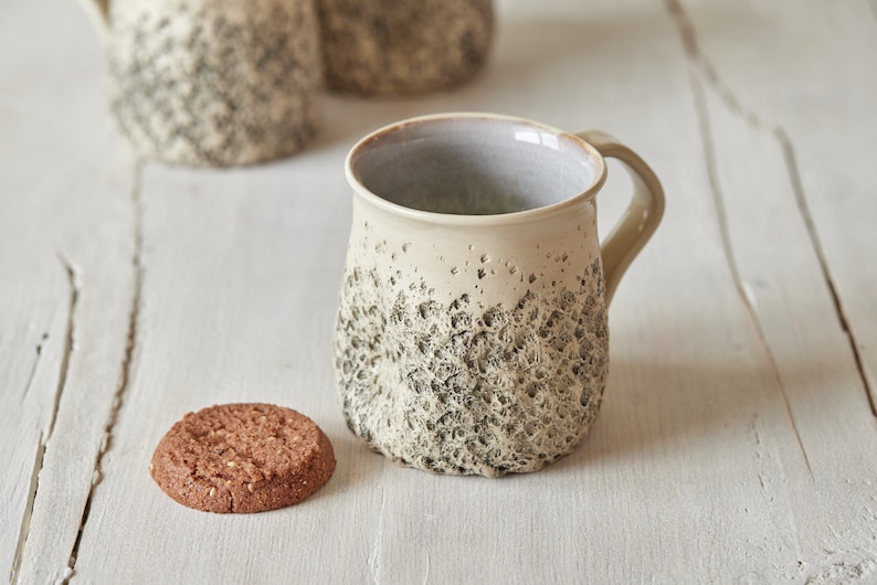 May include: A beige ceramic mug with a textured, speckled surface and a light gray interior. The mug has a rounded body and a handle. A chocolate chip cookie is sitting on a white wooden surface next to the mug.