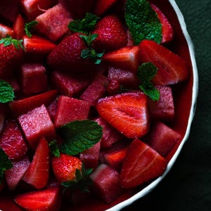 Puede incluir: Un cuenco lleno de una vibrante ensalada de frutas compuesta por cubos de sandía roja y fresas frescas cortadas por la mitad, adornada con hojas de menta de color verde brillante. El cuenco tiene un interior rojo intenso y un borde blanco.