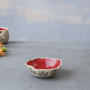 May include: A set of four small, red ceramic bowls with a textured, white rim. The bowls are arranged on a gray surface.