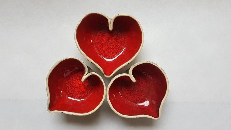 May include: Three heart-shaped ceramic bowls in a red glaze with a white rim. The bowls are arranged in a triangular pattern.