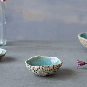 May include: Handmade ceramic bowls with a textured, speckled exterior and a bright turquoise interior. The bowls have an organic, irregular shape. The image also includes clear glass bottles and purple flowers, all set against a grey background.