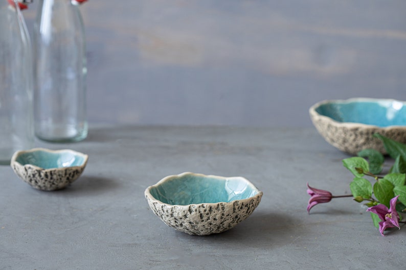 May include: Handmade ceramic bowls with a textured, speckled exterior and a bright turquoise interior. The bowls have an organic, irregular shape. The image also includes clear glass bottles and purple flowers, all set against a grey background.