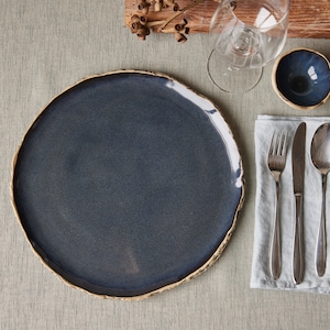 May include: A dark blue ceramic dinner plate with a slightly uneven rim sits on a light gray tablecloth. A white linen napkin with a silver fork, knife, and spoon is to the right of the plate. A small blue ceramic bowl is to the right of the napkin.