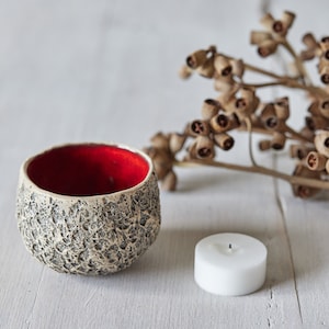 May include: A white ceramic candle holder with a textured surface and a red interior. The candle holder is next to a white pillar candle and a sprig of dried eucalyptus.