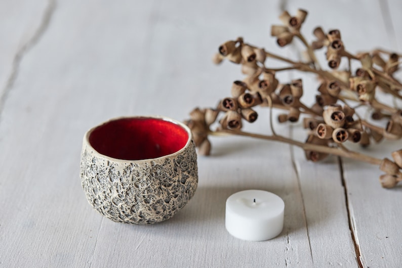 May include: A white ceramic candle holder with a textured surface and a red interior. The candle holder is next to a white pillar candle and a sprig of dried eucalyptus.