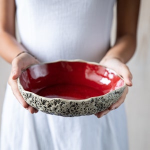 May include: A red ceramic bowl with a textured, speckled rim. The bowl is held by a person's hands.