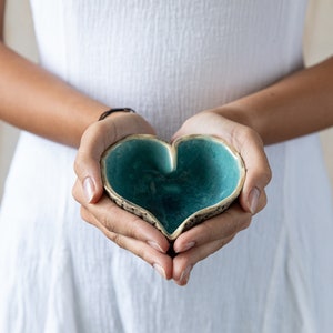 Puede incluir: Una persona sosteniendo un cuenco de cer&aacute;mica en forma de coraz&oacute;n con un esmalte turquesa. El cuenco tiene un exterior texturizado y un interior liso.