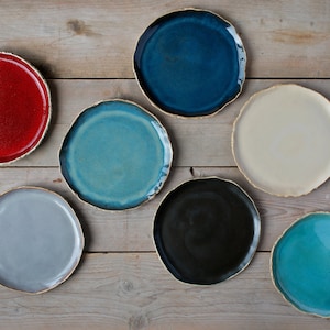 May include: Eight ceramic plates in various colors, including red, blue, gray, white, and turquoise, with a gold rim. The plates are arranged in a row on a wooden surface.