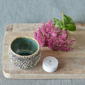 May include: A small, teal and brown speckled ceramic bowl sits on a wooden cutting board with a white candle and a bouquet of pink flowers.
