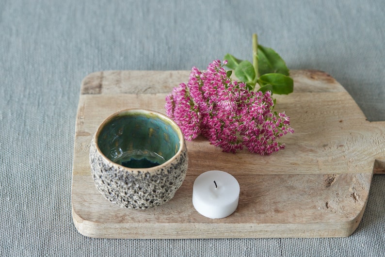 May include: A small, teal and brown speckled ceramic bowl sits on a wooden cutting board with a white candle and a bouquet of pink flowers.