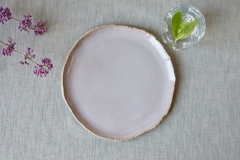 May include: A round ceramic plate with a smooth, light lavender-gray glaze and an uneven, textured, natural-looking rim. The plate is positioned next to a small sprig of purple berries and a clear glass bowl containing water and green mint leaves.