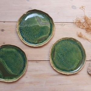 May include: Three round ceramic plates with irregular, wavy edges. The plates feature a glossy, mottled glaze in shades of forest green, emerald, and olive, with a textured, crystalline surface pattern and smooth, dark green solid sections.
