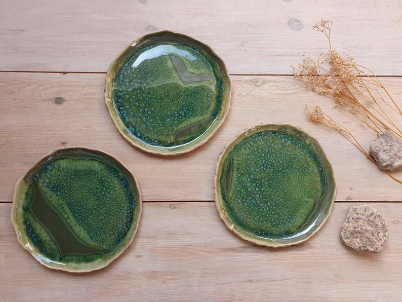 May include: Three round ceramic plates with irregular, wavy edges. The plates feature a glossy, mottled glaze in shades of forest green, emerald, and olive, with a textured, crystalline surface pattern and smooth, dark green solid sections.