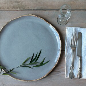 May include: A gray ceramic plate with a sprig of green leaves on a wooden table. A white linen napkin with a silver fork and knife is to the right of the plate. A clear glass is to the upper right of the plate.
