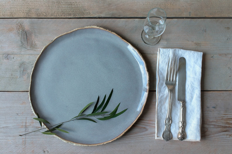 May include: A gray ceramic plate with a sprig of green leaves on a wooden table. A white linen napkin with a silver fork and knife is to the right of the plate. A clear glass is to the upper right of the plate.