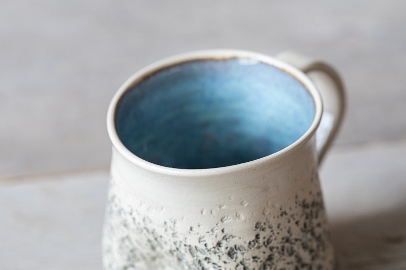 May include: A white ceramic mug with a speckled glaze and a light blue interior. The mug has a rounded shape and a handle.