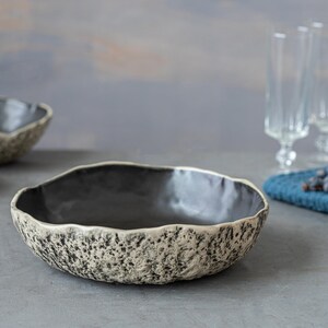 May include: Three ceramic bowls with a speckled, textured finish. The bowls are black on the inside and have a light gray exterior. The largest bowl is in the foreground and the two smaller bowls are behind it.