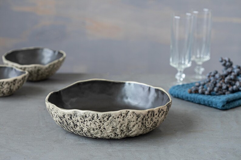 May include: Three ceramic bowls with a speckled, textured finish. The bowls are black on the inside and have a light gray exterior. The largest bowl is in the foreground and the two smaller bowls are behind it.