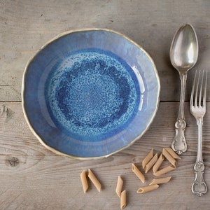 May include: A blue ceramic bowl with a white and blue speckled glaze sits on a wooden surface with a silver spoon and fork and scattered pieces of pasta.