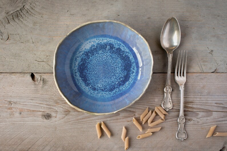 May include: A blue ceramic bowl with a white and blue speckled glaze sits on a wooden surface with a silver spoon and fork and scattered pieces of pasta.