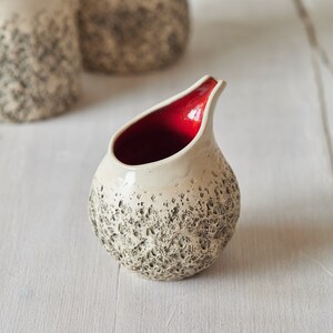 Puede incluir: Un peque&ntilde;o creamer de cer&aacute;mica blanca con una superficie texturizada y un interior rojo. El creamer tiene un cuerpo redondeado y un pico.