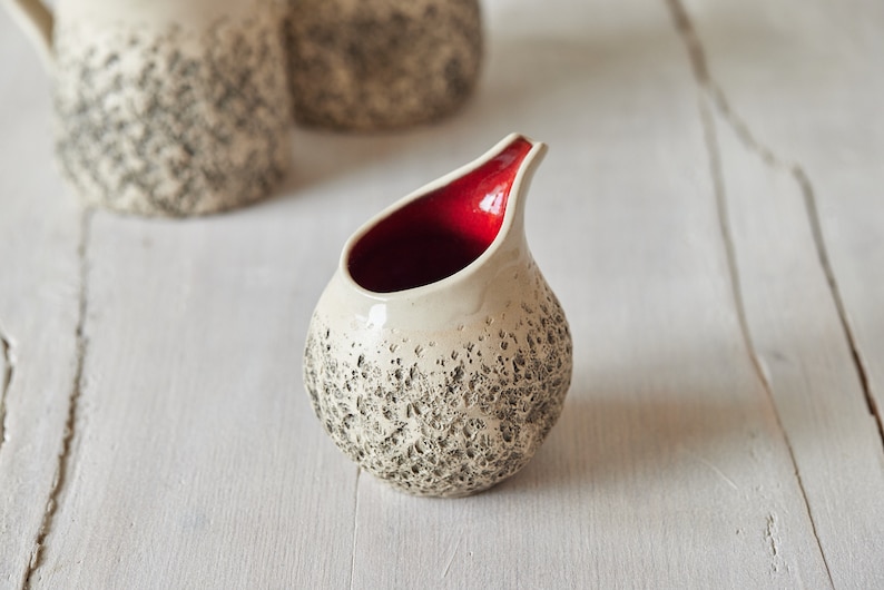 Puede incluir: Un peque&ntilde;o creamer de cer&aacute;mica blanca con una superficie texturizada y un interior rojo. El creamer tiene un cuerpo redondeado y un pico.