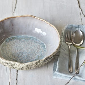 May include: A light blue and gray ceramic bowl with a textured rim. The bowl is sitting on a white wooden surface. A blue linen napkin with silver cutlery and a sprig of olive branches is in the background.