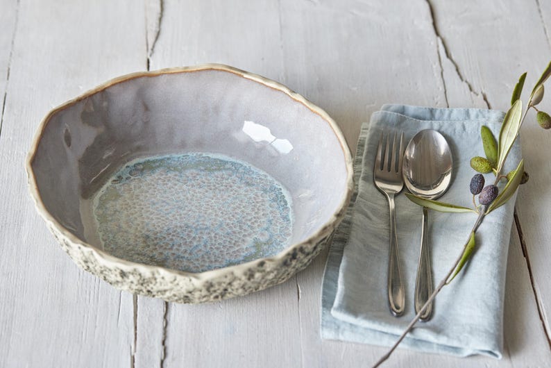 May include: A light blue and gray ceramic bowl with a textured rim. The bowl is sitting on a white wooden surface. A blue linen napkin with silver cutlery and a sprig of olive branches is in the background.