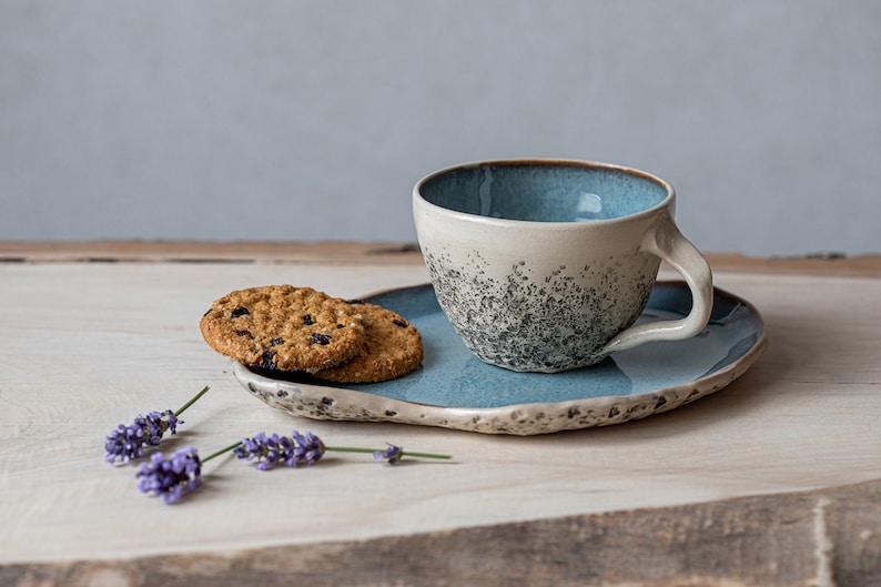May include: A blue and white ceramic teacup and saucer with a speckled glaze. The saucer has a wavy edge and is sitting on a wooden surface with two oatmeal cookies and lavender sprigs.
