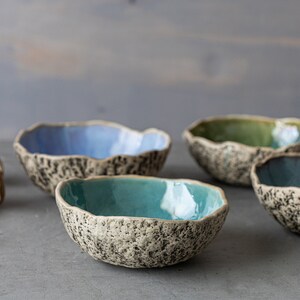 May include: Four small ceramic bowls with speckled exteriors and smooth, colorful interiors. The bowls are arranged in a row on a gray surface. The bowls are blue, green, teal, and light blue.