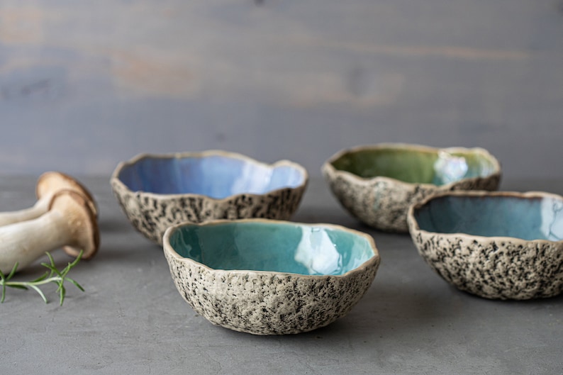 May include: Four small ceramic bowls with speckled exteriors and smooth, colorful interiors. The bowls are arranged in a row on a gray surface. The bowls are blue, green, teal, and light blue.