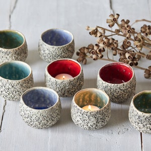 May include: Eight small ceramic candle holders with speckled white glaze. The holders are in various colors, including red, blue, and green. Some of the holders have tealight candles inside.