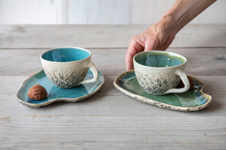May include: Two ceramic teacups with speckled glaze and matching saucers. The cups are teal and green with white interiors. One saucer has a chocolate truffle on it.