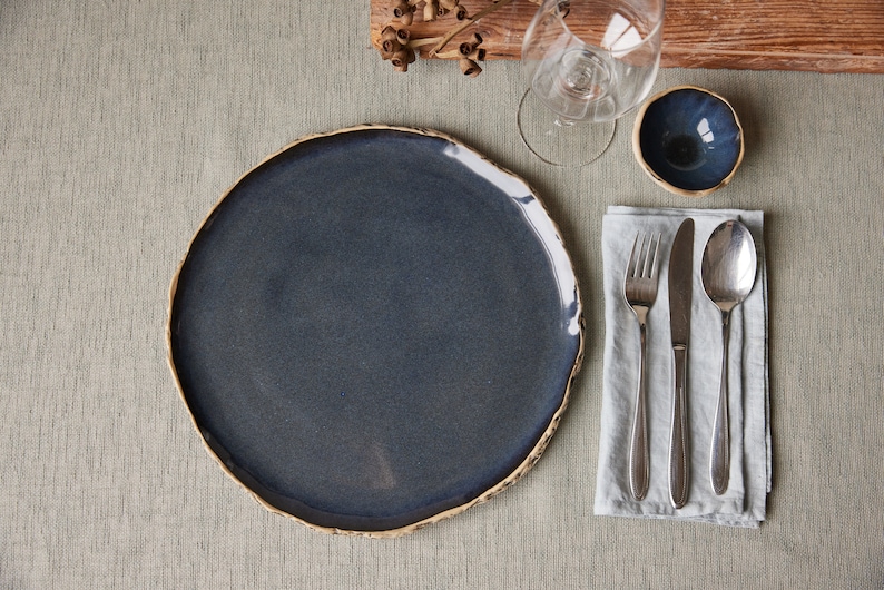 May include: A dark blue ceramic dinner plate with a slightly uneven rim sits on a light gray tablecloth. A white linen napkin with a silver fork, knife, and spoon is to the right of the plate. A small blue ceramic bowl is to the right of the napkin.