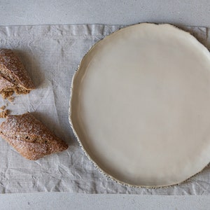 May include: A white ceramic plate with a slightly irregular rim sits on a beige linen placemat. Two halves of a brown bread roll are on the placemat.