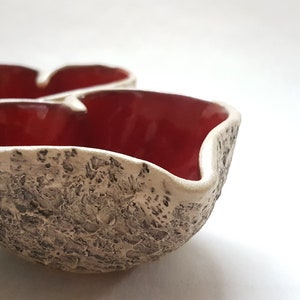 May include: Three small ceramic bowls with a textured exterior and a red glaze on the inside. The bowls are shaped like hearts and are arranged in a cluster.