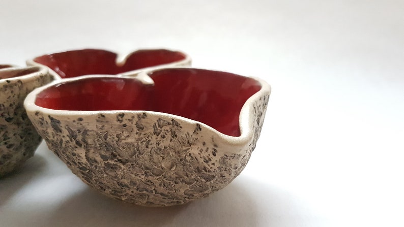 May include: Three small ceramic bowls with a textured exterior and a red glaze on the inside. The bowls are shaped like hearts and are arranged in a cluster.
