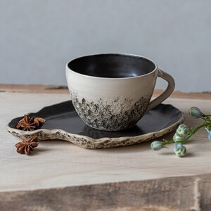 May include: A white ceramic coffee cup with a black interior and black speckles on the exterior, sitting on a black ceramic saucer with a wavy edge. The cup and saucer are on a light brown wooden surface with star anise pods and a green stem with small blue flowers.