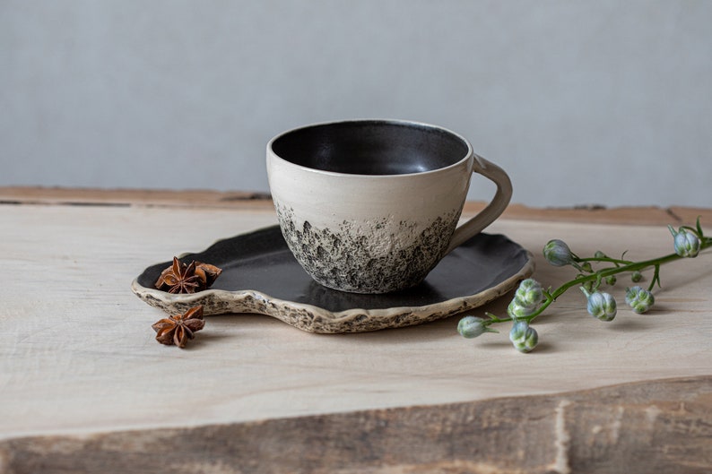 May include: A white ceramic coffee cup with a black interior and black speckles on the exterior, sitting on a black ceramic saucer with a wavy edge. The cup and saucer are on a light brown wooden surface with star anise pods and a green stem with small blue flowers.