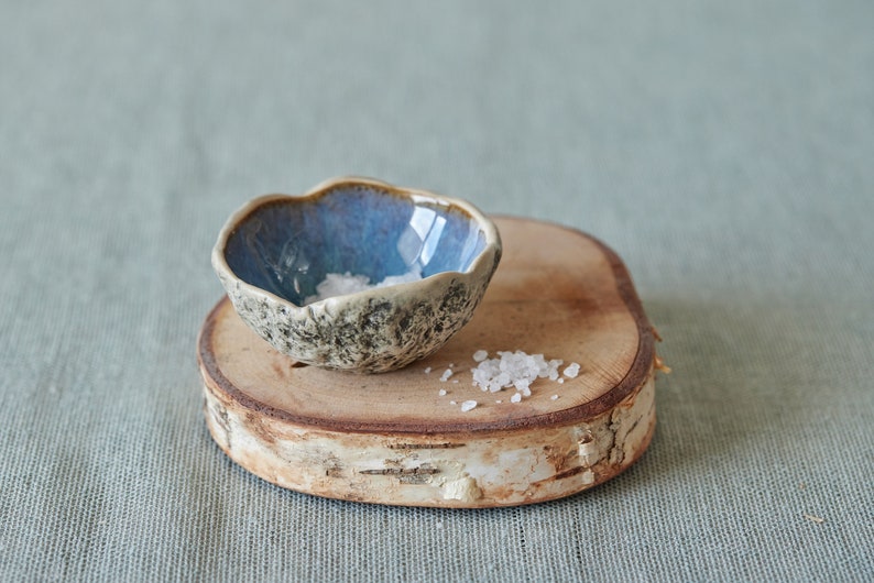 May include: A small, blue and white ceramic bowl filled with salt sits on a wooden slice. The bowl has a rough, textured exterior.