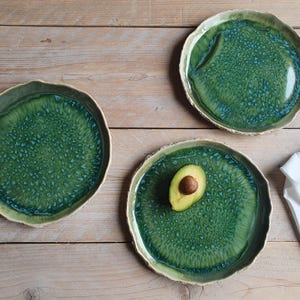 May include: Three round ceramic plates with irregular, wavy edges featuring a glossy green glaze with a textured, mottled blue pattern. One plate holds a halved avocado with a visible pit, and a silver fork and knife rest on a white napkin.