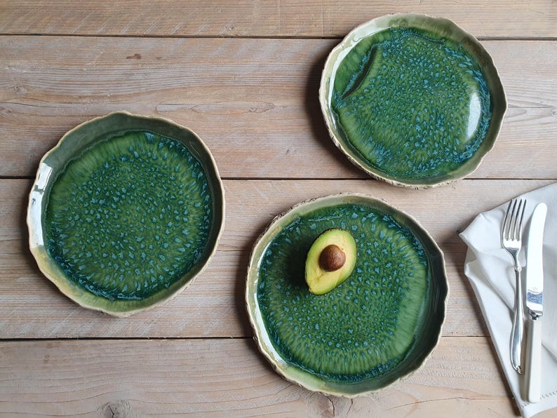 May include: Three round ceramic plates with irregular, wavy edges featuring a glossy green glaze with a textured, mottled blue pattern. One plate holds a halved avocado with a visible pit, and a silver fork and knife rest on a white napkin.