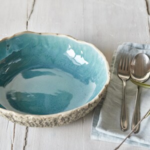 May include: A teal blue ceramic bowl with a textured rim sits on a white wooden surface. A blue linen napkin with silver cutlery and a sprig of olive branches is to the right of the bowl.