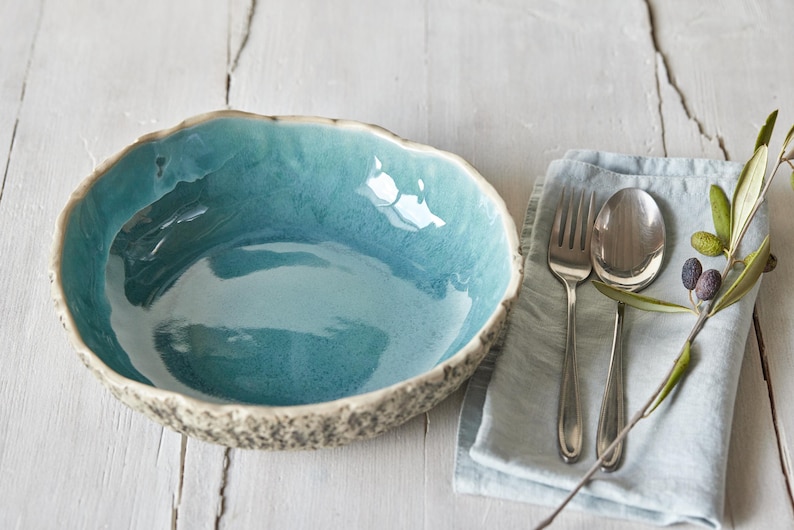 May include: A teal blue ceramic bowl with a textured rim sits on a white wooden surface. A blue linen napkin with silver cutlery and a sprig of olive branches is to the right of the bowl.