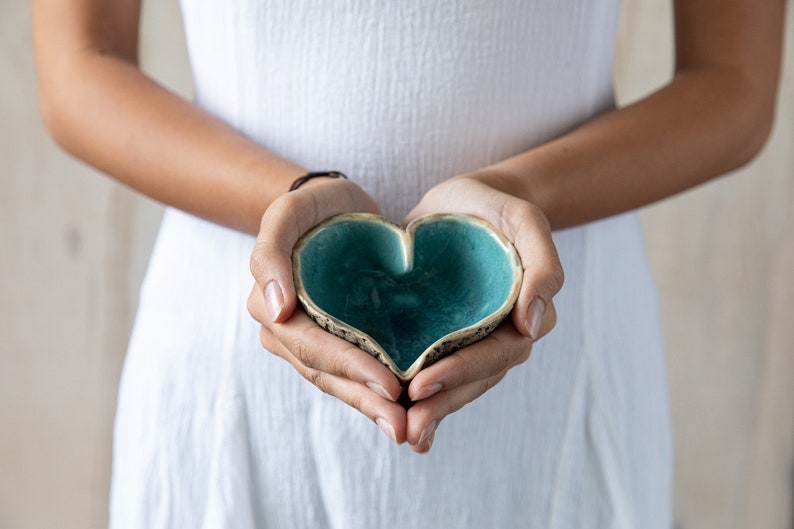 May include: A person holding a heart-shaped ceramic bowl with a teal glaze. The bowl has a rough, textured exterior.