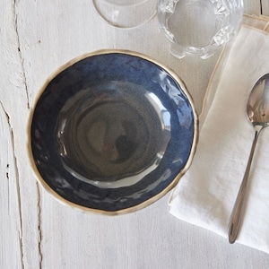 Könnte beinhalten: Eine dunkelblaue Keramikschüssel mit leicht gewelltem Rand. Die Schüssel ist leer und steht auf einer weißen Holzoberfläche.