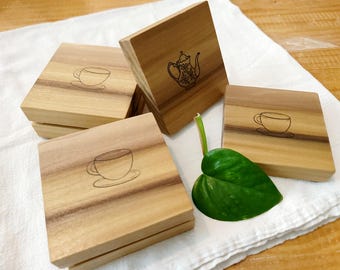 May include: Four wooden coasters with different designs. One coaster features a teapot, while the other three feature a cup and saucer. The coasters are on a white surface with a green leaf.