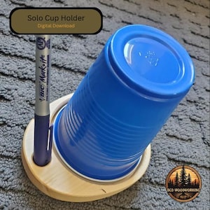 May include: A wooden solo cup holder with a blue plastic cup and a purple pen. The holder has a circular base with a hole for the pen. The text "Solo Cup Holder Digital Download" is in a gold box.